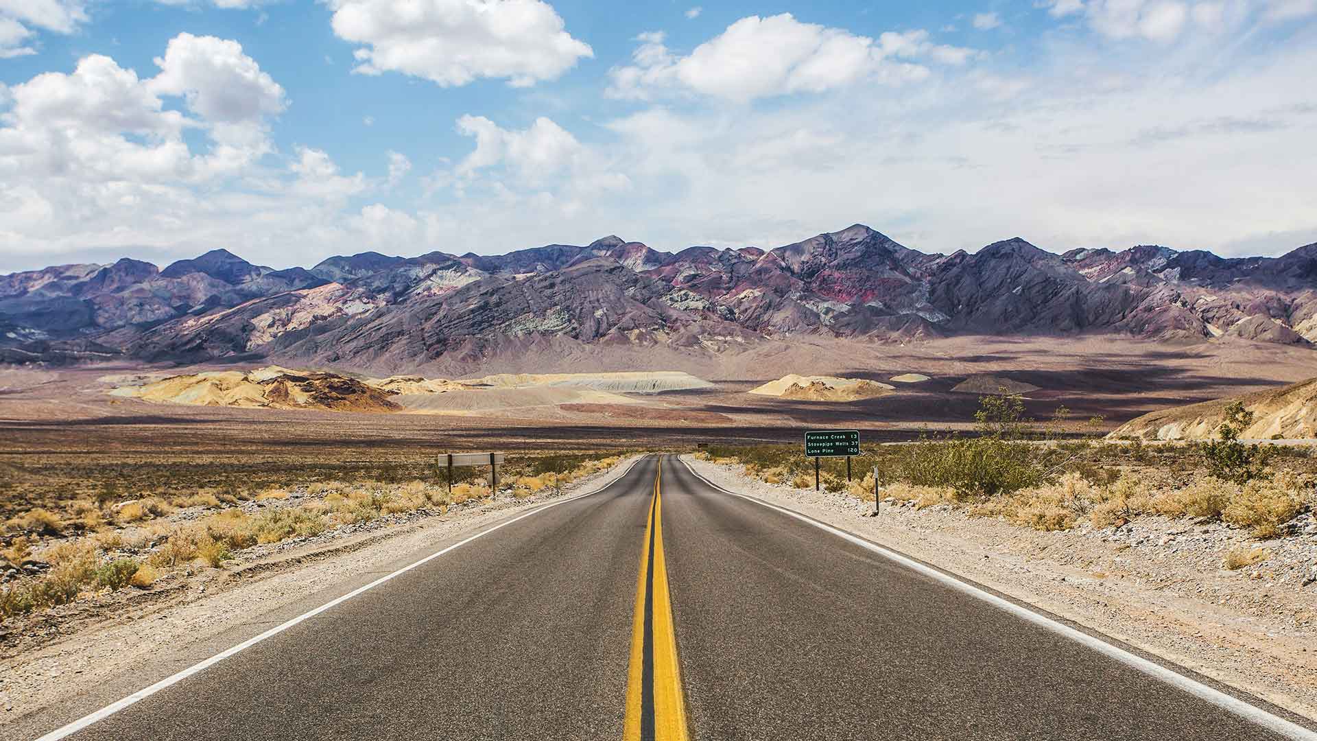 A straight desert highway leads into multicolored mountains under a blue sky with scattered clouds, capturing the essence of Route 66—one of the most iconic road trips in the USA.
