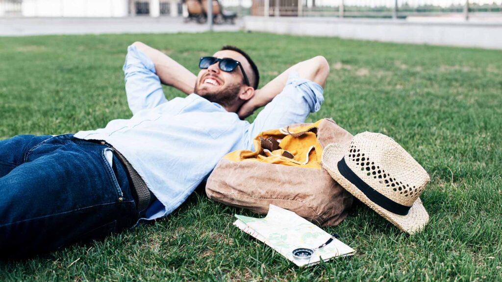 Man lying on grass with a backpack and hat, showcasing simple travel hacks for a stress-free trip