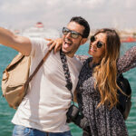 Young couple taking a fun travel selfie by the sea, excited to explore with Zee Business travel deals