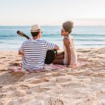 A couple enjoys a peaceful moment on a sandy beach at sunset, with one playing guitar and the other smiling—capturing the essence of affordable beach vacations filled with simple joys and relaxation.