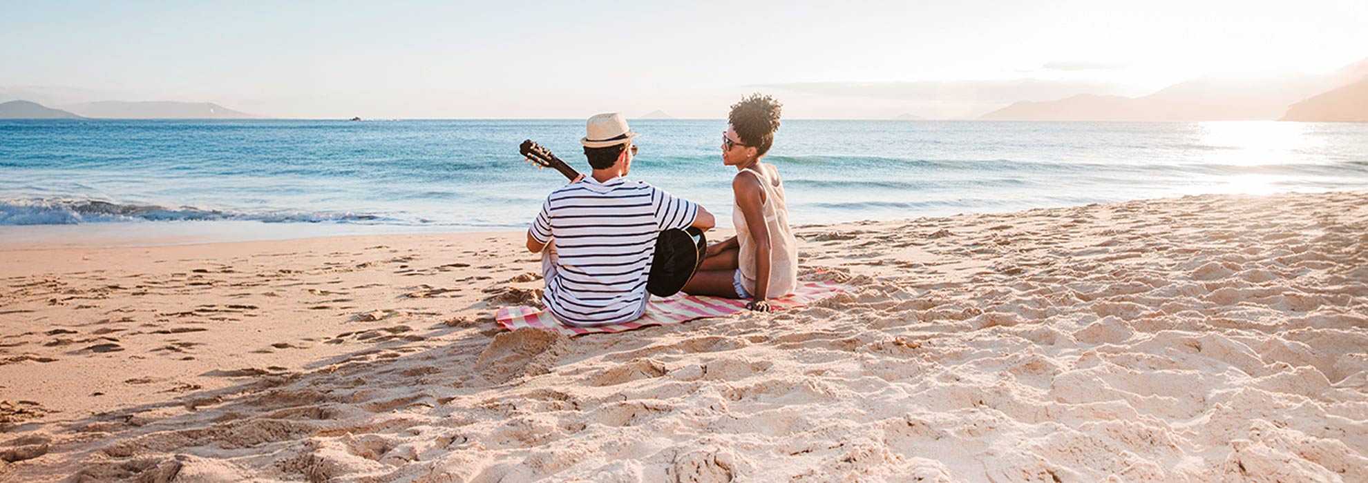 A couple enjoys a peaceful moment on a sandy beach at sunset, with one playing guitar and the other smiling—capturing the essence of affordable beach vacations filled with simple joys and relaxation.