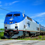 Modern Amtrak passenger train with a blue and silver locomotive traveling through lush greenery on a clear day.
