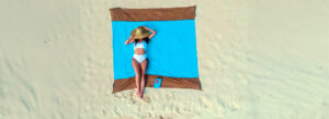 A woman relaxing on a beach towel, wearing a white swimsuit and a wide-brimmed hat, enjoying the serene beach setting.